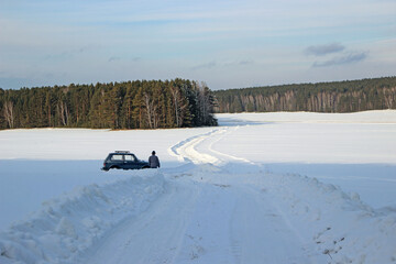 The snow-covered winding road disappears into the forest. The man near the car looks into the distance.