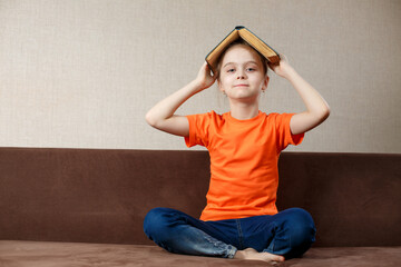 Happy little schoolgirl with book on her head, wearing orange t-shirt and jeans , sitting in sofa at home