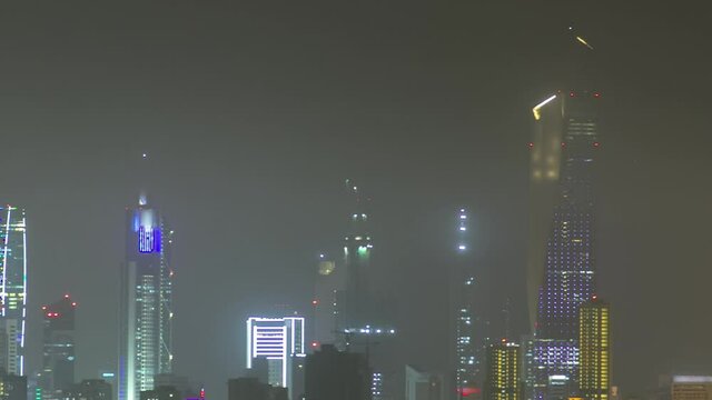 Skyline with Skyscrapers night timelapse in Kuwait City downtown illuminated at dusk. View from beach with reflection in water at foggy weather. Kuwait City, Middle East.