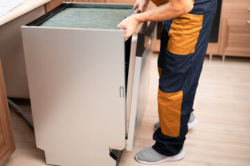 A man or service worker in special clothing installs, disassembles or tries on a built-in dishwasher in a niche of kitchen furniture.