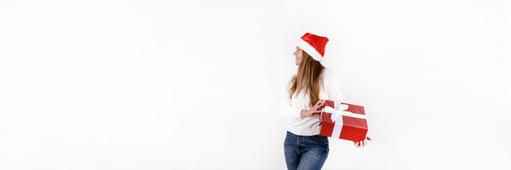 Young beautiful woman in red santa hat and white sweater holding gift box and looking to the side isolated on white background. Banner size, empty space