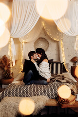 Fototapeta na wymiar Beautiful couple sitting on the bed near decorated Christmas tree, smiling. Charming husband cuddle adorable wife, enjoy tenderness. Handsome man celebrate xmas eve with gorgeous woman at home