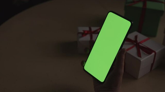 Handheld slow motion shot of mid aged female hands holding smartphone with green screen over wood vintage table with presents on background and dim window light