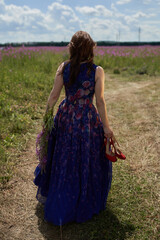 Fototapeta premium Young woman walking flowering field, turned away, gently touch yellow flowers. Brunette girl enjoys spring aromas, harmony. Backdrop dark clouds blue sky. Long hair fluttering wind. Denim sundress hat