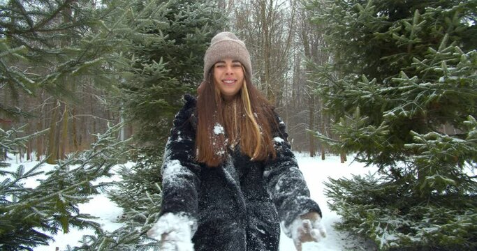 Playful young woman throws snow up and smiling in front of the tree in forest. First Snow And Winter Concept