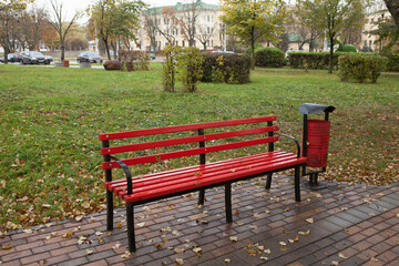 Red bench and red trash can in the autumn city park. There is nobody on the street.