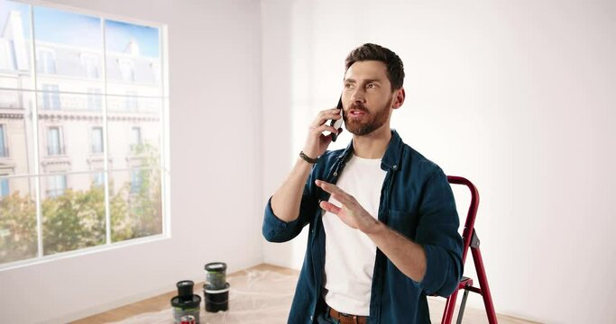 Handsome young joyful Caucasian handyman speaking on mobile phone with client in new apartment. Happy male chatting on smartphone during home repair and renovation. Redesigning room concept