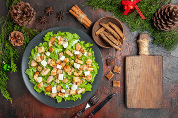 top view caesar salad on oval plate pine tree branches fork knife anises bowl with dried crust chopping board on dark red background