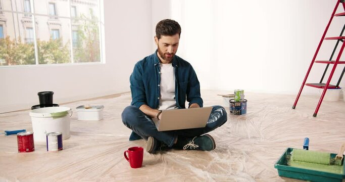 Caucasian happy bearded young handsome man sitting on floor in empty apartment during repair works texting and tapping on laptop computer. Renovation and improvement concept. Redesigning house concept