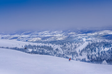 View of ski slope in Colorado ski resort with small figure of skier in red jacket on cold winter day; snow capped mountains, forest and silhouettes of buildings in background; cloud on mountain top