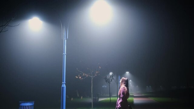 woman at night in the park approaches a bright lantern and looks up, thick fog, cold night, autumn, blue lamppost. Medium plan