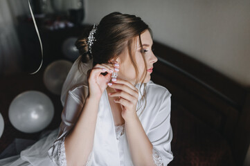 A beautiful bride in a white robe with curly hair sits on the bed and straightens an earring on her ear. Wedding portrait.