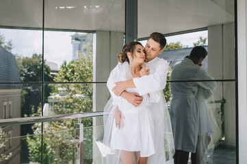 Naklejka premium The bride and groom in white robes embrace tenderly against the background of a mirrored wall in an expensive hotel. Morning of the newlyweds.