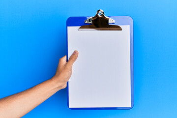 Young hispanic hand holding clipboard over isolated blue background.