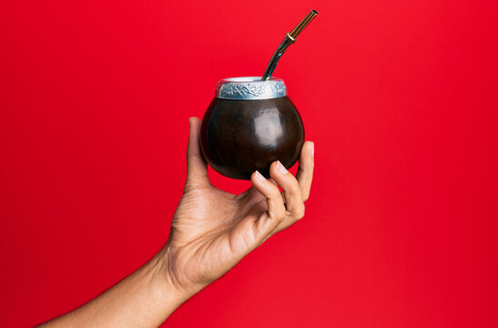 Hand of hispanic man holding cup of mate infusion beverage over isolated red background.