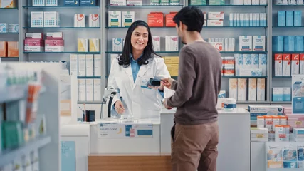Photo sur Aluminium Pharmacie Pharmacie Drugstore : l& 39 homme choisit un médicament, vient au comptoir, parle à une belle pharmacienne caissière, paie les produits de santé à la caisse à l& 39 aide d& 39 une carte de crédit pour paiement sans contact