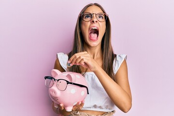 Young hispanic woman holding piggy bank with glasses and coin angry and mad screaming frustrated...