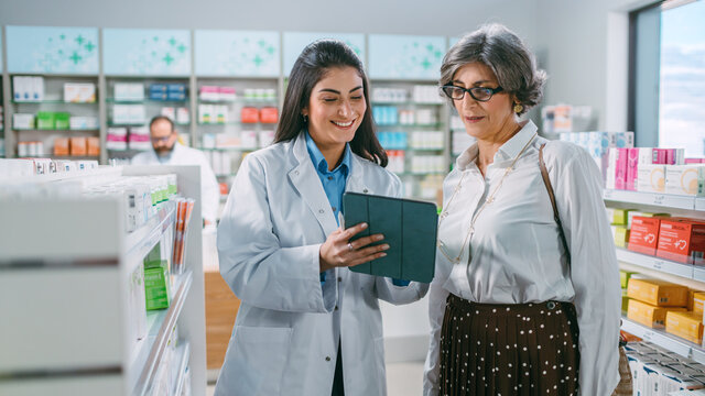 Pharmacy Drugstore: Senior Woman Chooses to Buy Medicins. Professional, Helpful Pharmacist Uses Digital Tablet Computer and Advicing Customer Best Option. Modern Pharma Store Health Care Products