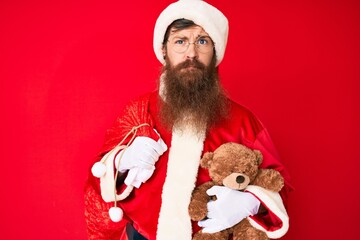 Handsome young red head man with long beard wearing santa claus costume holding teddy bear skeptic and nervous, frowning upset because of problem. negative person.