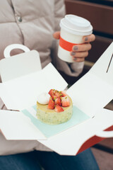 Close-up of woman hang holding cup of take away coffee and going to eat pistachio pastry flavored with fresh strawberries gluten free and vegan in box to go. Natural light, selective focus photo