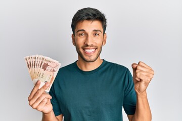 Young handsome man holding south korean won banknotes screaming proud, celebrating victory and...