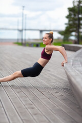 Young woman in sportswear does push-ups from bench on wooden embankment.
