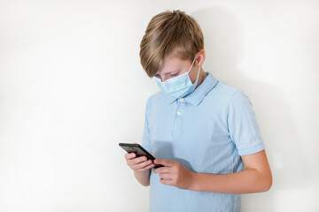 boy in protective face mask plays on smartphone in online game. schoolboy watches homework on phone during coronavirus quarantine