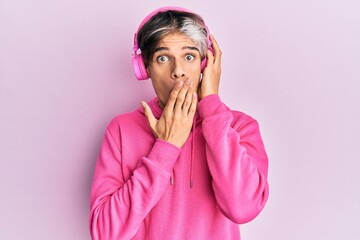 Young hispanic man listening to music using headphones covering mouth with hand, shocked and afraid...