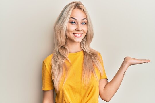 Beautiful caucasian blonde girl wearing casual tshirt smiling cheerful presenting and pointing with palm of hand looking at the camera.