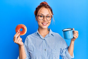 Young redhead woman eating breakfast holding chocolate donut and coffee smiling with a happy and...