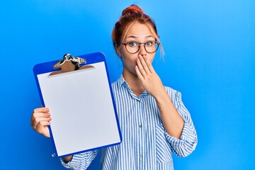 Young redhead woman holding clipboard with blank space covering mouth with hand, shocked and afraid...