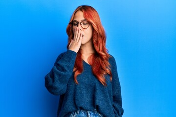 Young beautiful redhead woman wearing casual sweater and glasses over blue background bored yawning tired covering mouth with hand. restless and sleepiness.