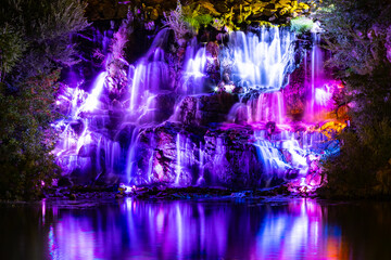 Waterfall in neon color. Flowing water illuminated by multicolored light. Night landscape with a colorful waterfall near the river