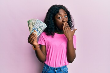 Beautiful african young woman holding dollars covering mouth with hand, shocked and afraid for...