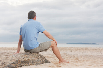 Mature man sitting on a beach looking at the sea with an island