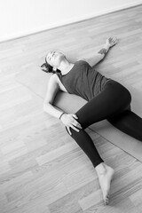 Naklejka premium A woman in her early 30's doing yoga on the floor, performing the spinal twist pose in her home interior. Black and white vertical format