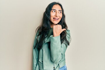 Hispanic teenager girl with dental braces wearing green leather jacket pointing thumb up to the...