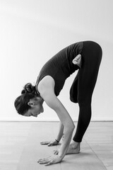 Side view of a young brunette woman on her way to down to a standing forward fold with lotus legs, forward bending asana or yoga pose.
