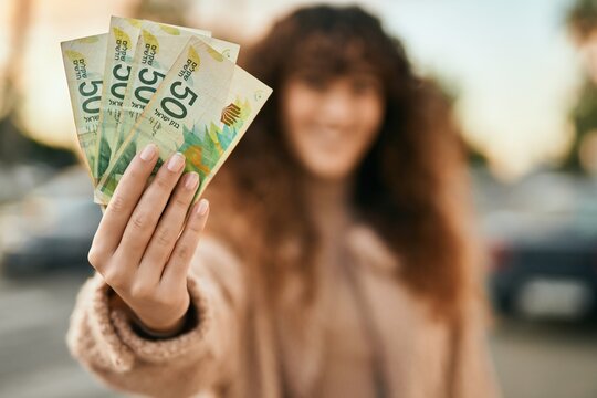 Young hispanic woman smiling happy holding israel shekels banknotes at the city