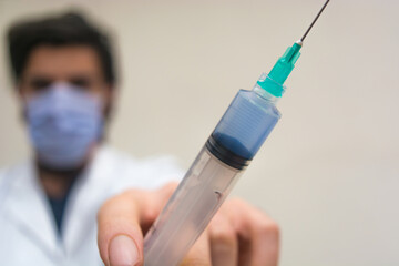 Disposable plastic syringe with blue liquid vaccine inside holding with fingers focused, with a doctor with white coat and mask unfocused in the background.
