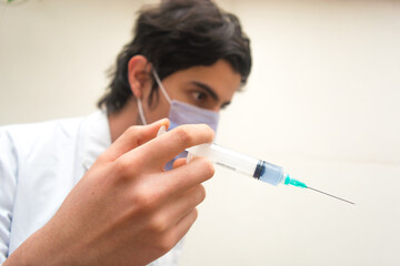 Disposable plastic syringe with blue liquid vaccine inside holding with fingers focused, with a doctor with white coat and mask unfocused in the background.