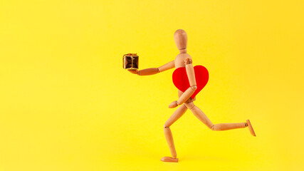 Fototapeta na wymiar A running wooden man carries a red heart and a gift in his hand on a yellow illuminating background.