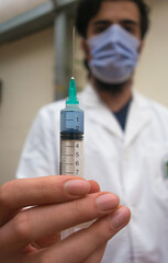 Disposable plastic syringe with blue liquid vaccine inside holding with fingers focused, with a doctor with white coat and mask unfocused in the background.