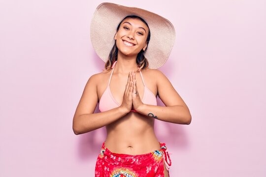 Young woman wearing bikini and hat praying with hands together asking for forgiveness smiling confident.