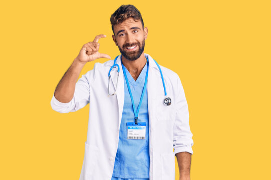 Young hispanic man wearing doctor uniform and stethoscope smiling and confident gesturing with hand doing small size sign with fingers looking and the camera. measure concept.