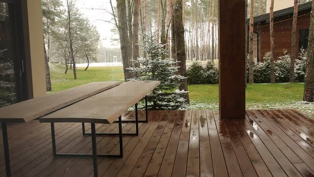 Wooden house in the forest, the first snow falls on the trees and grass. In the foreground is a wooden table on a wooden floor. In the background is a Christmas tree and pine trees.