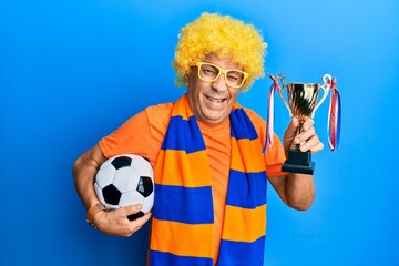 Senior hispanic man football hooligan cheering game holding ball and trophy winking looking at the camera with sexy expression, cheerful and happy face.