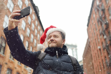Obraz premium Middle aged man tourist in Santa Claus hat takes selfie on street near the Manhattan Bridge in New York City on Christmas Eve. Winter holidays in NYC.