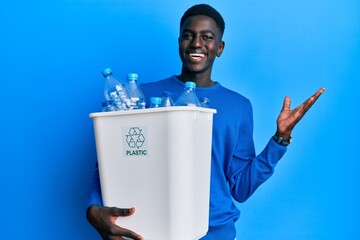 Young african american man holding recycling wastebasket with plastic bottles celebrating victory...
