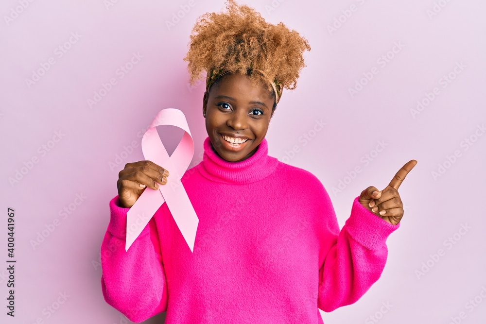 Canvas Prints Young african woman with afro hair holding pink cancer ribbon smiling happy pointing with hand and finger to the side